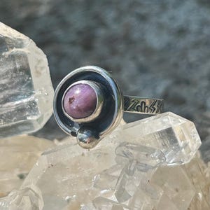 Puede incluir: Un anillo de plata de ley con una gema redonda de color púrpura claro engastada en un bisel circular. El anillo tiene una banda decorativa con diseños de rayos grabados. El anillo se exhibe sobre una superficie cristalina transparente.