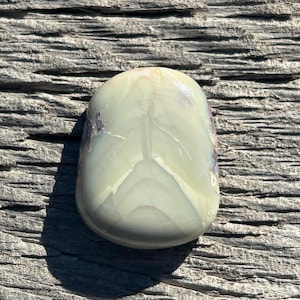 Könnte beinhalten: Ein glatter, oval geformter, hellgrüner Stein mit einem blattartigen Muster. Der Stein hat subtile violette und weiße Adern und liegt auf einer verwitterten, grauen Holzoberfläche. Der Stein ist ein natürlicher Edelstein.