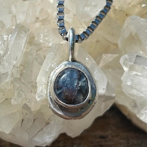 May include: A silver pendant necklace featuring a round, blue-grey stone. The stone is set in a textured silver bezel and hangs from a matching silver chain. The necklace is displayed against a backdrop of clear crystals.