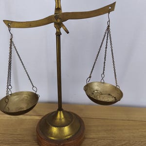 May include: A brass balance scale featuring a central vertical post with a decorative eagle topper, a horizontal beam, and two hanging pans suspended by thin metal chains. The entire structure is mounted on a circular wooden base.