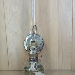 Könnte beinhalten: Eine Vintage-Öllampe mit einem transparenten Glasschornstein und einem runden, transparenten Glassockel. Die Lampe hat einen Metallbrenner und einen Docht, der im Inneren sichtbar ist. Die Lampe ist zum Aufhängen konzipiert.