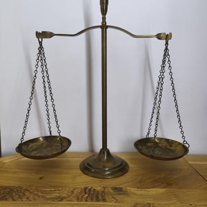 Peut inclure: Une balance à plateaux en laiton de style vintage composée d'un montant central vertical sur une base circulaire, d'un fléau horizontal et de deux coupelles circulaires peu profondes suspendues par de fines chaînes métalliques aux extrémités.