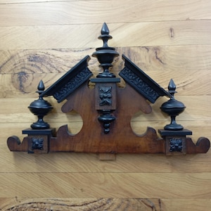 Könnte beinhalten: Dekorative Wandverzierung aus Holz mit dunkelbraunem Finish. Das Ornament hat ein zentrales Holzelement mit kunstvollen Schnitzereien, flankiert von zwei abgewinkelten, schwarzen, dekorativen Paneelen. Das Design umfasst Fialen und dekorative Elemente.