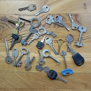 May include: A collection of various metal keys, in different shapes and sizes, scattered on a wooden surface. Some keys have black or blue plastic handles. The keys are in shades of silver, gold, and gray.