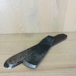 Könnte beinhalten: Zwei rostige Metallklingen mit abgerundeten Kanten, wahrscheinlich für die Holzbearbeitung verwendet, liegen gekreuzt auf einer hellen Holzoberfläche.