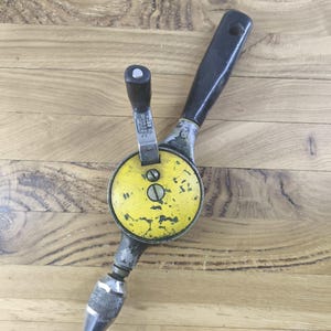 Op de afbeelding: Een vintage Stanley handboor met een gele ronde tandwielkast met zichtbare verfslijtage, een zwarte handgreep, een metalen zwengel met een zwarte knop en een gekartelde metalen boorkop aan de onderkant. Het gereedschap bevat markeringen die aangeven dat het in Engeland is gemaakt.