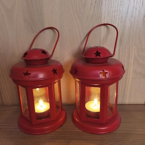 Op de afbeelding: Twee rode metalen lantaarns met een ronde voet en een handvat aan de bovenkant. Elke lantaarn heeft stervormige uitsparingen langs de bovenrand en koepel, met heldere glazen panelen rondom een brandend wit waxinelichtje in de centrale kamer.