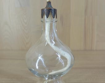 May include: A clear glass oil lamp with a bulbous body and a flared base. The lamp has a metal cap with a wick extending from the top. The wick is white, and the metal cap is dark. The lamp is designed to hold oil for burning.
