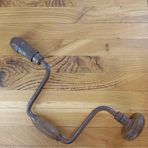 May include: A vintage hand drill with a wooden handle and a rusty, curved metal frame. The drill is resting on a light brown wooden surface, highlighting its antique design and construction.