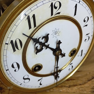 Peut inclure: Gros plan sur un cadran d'horloge antique avec un fond blanc et des chiffres noirs. L'horloge a une lunette dorée et des aiguilles noires ornées. Le cadran de l'horloge comporte un petit motif en forme d'éclat d'étoile avec les lettres "SJ" à l'intérieur.