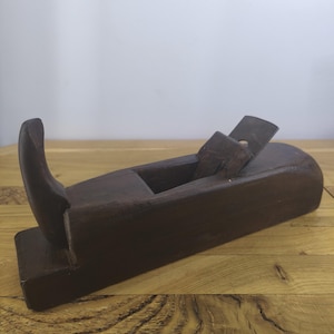 May include: A dark brown wooden hand plane featuring a curved handle at the rear and a metal blade set into the top centre of the rectangular body. The tool has a smooth, weathered finish and a distinct grain pattern visible across its surface.