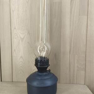 Könnte beinhalten: Eine Vintage-Öllampe mit einem transparenten Glaszylinder und einem dunkelblauen Sockel. Die Lampe hat einen runden Sockel, eine schwarze Metallbrenneranordnung und einen hohen, zylindrischen Glaszylinder. Die Lampe ist für Öl und einen Docht zur Beleuchtung konzipiert.