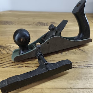 Könnte beinhalten: Ein Metall-Handhobel mit grünem Körper, einem dunklen Holzgriff und einem runden Holzknauf. Das Werkzeug verfügt über einen metallischen Klingenverstellmechanismus und wird von einem separaten schwarzen Metallanschlag mit Gewindestange begleitet.