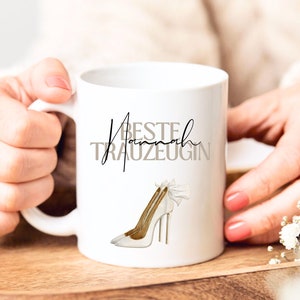 Puede incluir: Taza de cerámica blanca con texto negro que dice "Beste Hannah Trauzeugin" y un zapato de tacón alto blanco con tacón dorado y un lazo blanco.