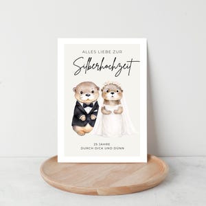 May include: A greeting card featuring two cartoon otters dressed for a wedding. One otter wears a tuxedo, the other a white dress and veil. The card has text in German, and is displayed on a wooden tray.