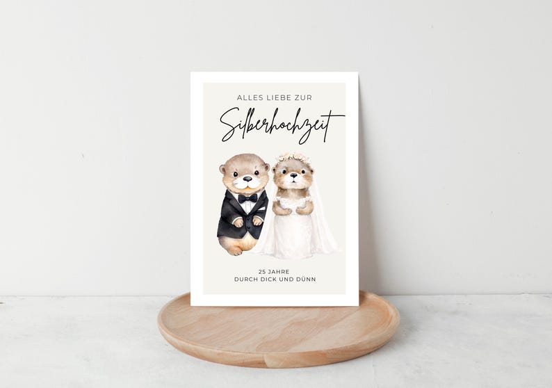 May include: A greeting card featuring two cartoon otters dressed for a wedding. One otter wears a tuxedo, the other a white dress and veil. The card has text in German, and is displayed on a wooden tray.