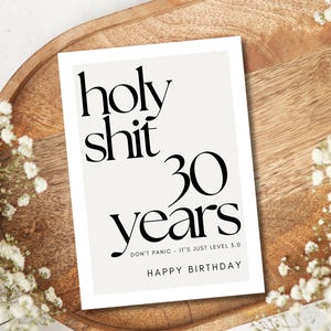Może przedstawiać: Biała kartka urodzinowa z czarnym tekstem. Na kartce napisano "holy shit 30 years", "Don't panic - it's just level 5.0" i "Happy Birthday". Kartka leży na drewnianej tacy, ozdobionej białymi kwiatami.