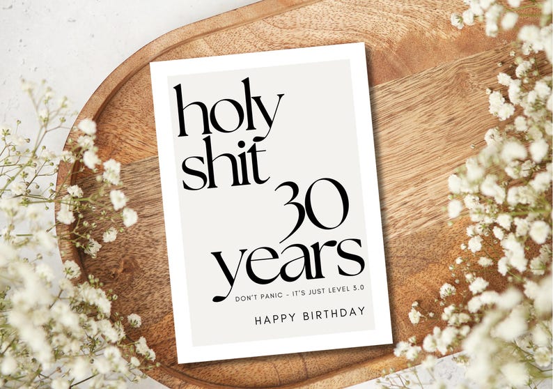 Może przedstawiać: Biała kartka urodzinowa z czarnym tekstem. Na kartce napisano "holy shit 30 years", "Don't panic - it's just level 5.0" i "Happy Birthday". Kartka leży na drewnianej tacy, ozdobionej białymi kwiatami.