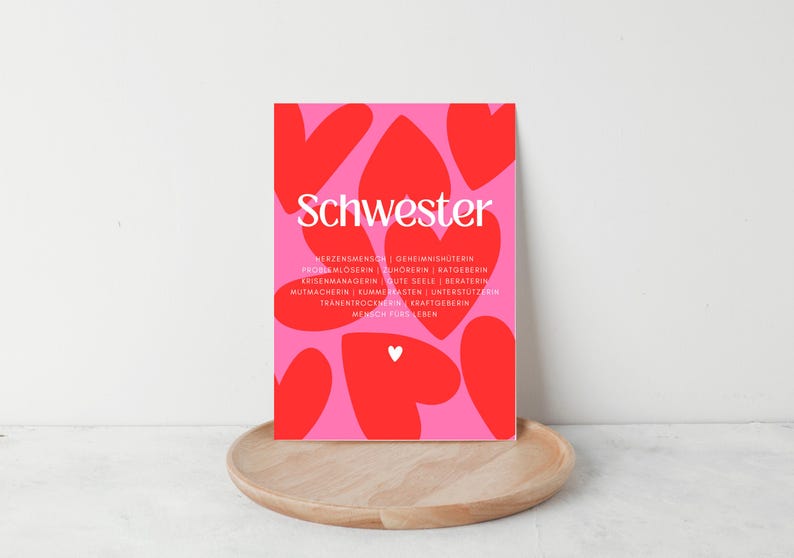 May include: A pink and red art print with the German word "Schwester" in white, surrounded by red heart shapes. The print includes additional text below the title. The print is displayed on a wooden tray.