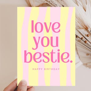 Könnte beinhalten: Eine Grußkarte mit gelb-rosa gestreiftem Hintergrund. Der Text "love you bestie." ist in Pink geschrieben, darunter steht "Happy Birthday". Die Karte wird von einer Hand gehalten.