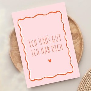 以下が含まれることがあります： オレンジ色の文字で「Ich hab's gut, ich hab dich」と書かれたピンク色のグリーティングカード。オレンジと白の波状のボーダーで囲まれています。小さなオレンジ色のハートが下にあります。カードは木製のプレートの上にあります。