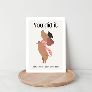May include: A graduation greeting card featuring a cartoon dachshund wearing a graduation cap and a pink scarf. The text "You did it" is at the top, with "HERZLICHEN GL&Uuml;CKWUNSCH" below. The card is on a wooden tray.