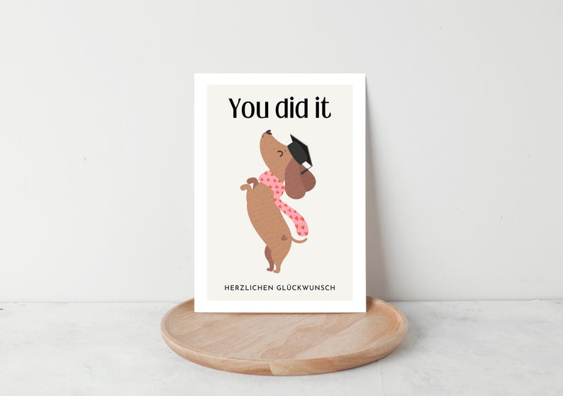 May include: A graduation greeting card featuring a cartoon dachshund wearing a graduation cap and a pink scarf. The text "You did it" is at the top, with "HERZLICHEN GL&Uuml;CKWUNSCH" below. The card is on a wooden tray.