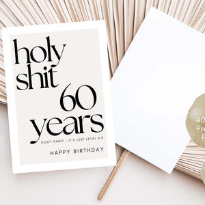 May include: A birthday card with the text "holy shit 60 years" in black font. The card also includes the phrases "DON'T PANIC - IT'S JUST LEVEL 6.0" and "HAPPY BIRTHDAY". A blank card and two circular labels are also visible.