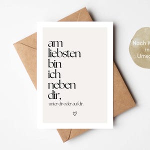 May include: A greeting card with the text "am liebsten bin ich neben dir, unter dir oder auf dir." in black font on a white background. The card is placed on a brown envelope. A small, round sticker with text is also visible.