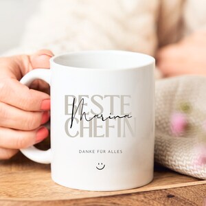 May include: White ceramic mug with the text "Beste Marina Chefin" in gray and black lettering. The text "Danke fur alles" is printed below the main text, and a smiley face is printed below that.