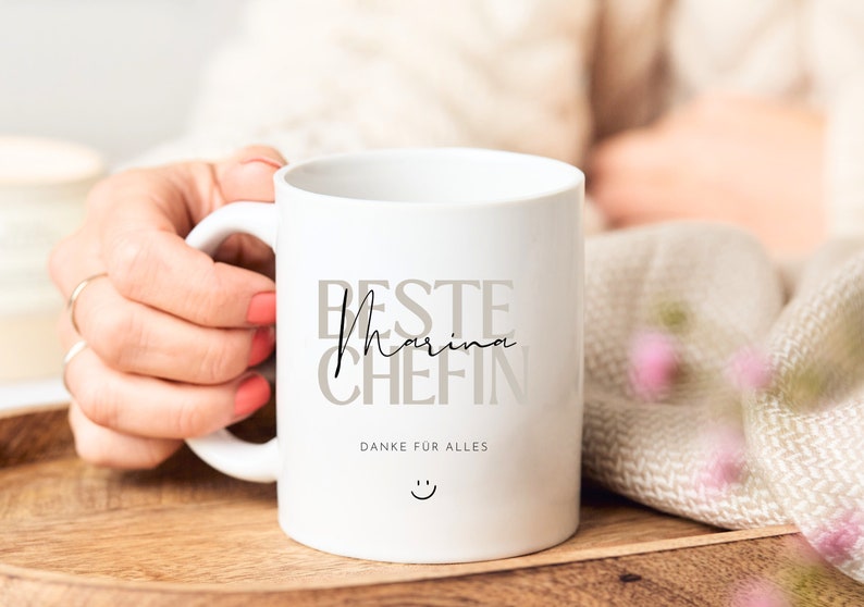 May include: White ceramic mug with the text "Beste Marina Chefin" in gray and black lettering. The text "Danke fur alles" is printed below the main text, and a smiley face is printed below that.