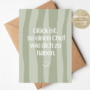 May include: A rectangular greeting card with a sage green background featuring vertical wavy lines in a darker shade. The text reads "Glück ist, so einen Chef wie dich zu haben." with a small white smiley face icon below the text. A brown envelope is visible.