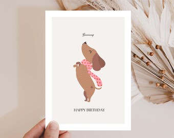 Puede incluir: Tarjeta de felicitación con un dibujo de un perro salchicha de dibujos animados con una bufanda rosa con corazones blancos. El perro está de pie sobre sus patas traseras, mirando hacia arriba. La palabra "Boooof" está encima del perro, y "HAPPY BIRTHAY" está debajo.