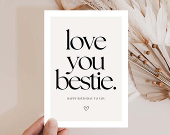 May include: A greeting card with the words "love you bestie" in black, stylized font. Below, the text "Happy Birthday to You" and a heart symbol are printed. The card is held by a hand, with dried flowers in the background.