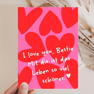 May include: A pink greeting card with red heart shapes. The card has the text "I love you, Bestie. Mit dir ist das Leben so viel schöner." in white. A small white heart is at the bottom. The card is held by a hand.