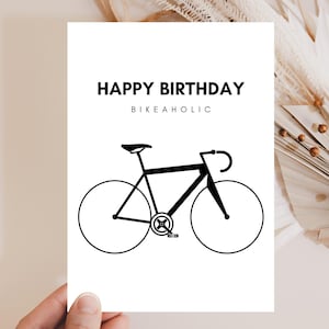 Könnte beinhalten: Eine weiße rechteckige Grußkarte mit dem schwarzen Text HAPPY BIRTHDAY und BIKEAHOLIC, der oben zentriert ist. Unter dem Text befindet sich eine minimalistische schwarze Strichzeichnung eines Rennrads mit zwei großen Rädern, Rahmen und Pedalen.