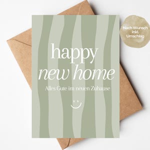 Könnte beinhalten: Eine salbeigrüne Grußkarte mit vertikalen Wellenstreifen, die den weißen Text "happy new home" und "Alles Gute im neuen Zuhause" sowie ein kleines weißes Smiley-Symbol zeigt, platziert auf einem braunen Papierumschlag mit einem runden Aufkleber.