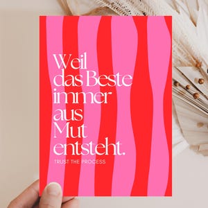 Peut inclure: Une carte rectangulaire avec un motif à rayures ondulées rouges et roses. La carte présente un texte blanc en allemand qui se lit "Weil das Beste immer aus Mut entsteht. TRUST THE PROCESS."