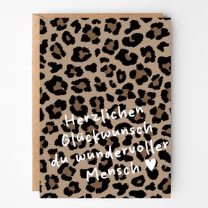 Puede incluir: Una tarjeta de felicitación rectangular con un patrón de estampado de leopardo en marrón y negro. La tarjeta muestra un texto en blanco que dice: Herzlichen Glückwunsch du wundervoller Mensch, seguido de un pequeño corazón blanco. Se ve un sobre marrón.
