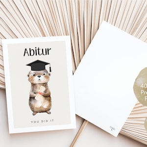 May include: A graduation card featuring a cartoon otter wearing a graduation cap and holding a diploma. The card is beige with black text that reads "Abitur" and "You Did It". The card is printed on 400g/m2 premium paper and is a DIN A6 postcard.