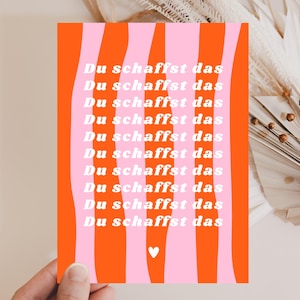 Peut inclure: Une affiche rectangulaire aux couleurs vives. Le fond présente des rayures verticales orange et rose. Le texte blanc "Du schaffst das" est répété plusieurs fois. Un petit cœur blanc est en bas.