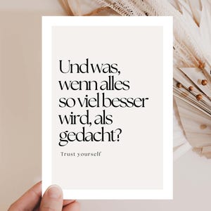 Puede incluir: Una impresión rectangular con el texto "Und was, wenn alles so viel besser wird, als gedacht?" y "Trust yourself" en fuente negra sobre un fondo crema. La impresión está sostenida por una mano, con flores secas en el fondo.