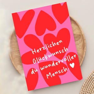 以下が含まれることがあります： 赤いハートの形とドイツ語のテキスト「Herzlichen Glückwunsch du wundervoller Mensch」が書かれたピンク色のグリーティングカード。小さな白いハートも付いています。カードは木製のプレートの上にあります。