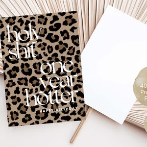 Puede incluir: Una tarjeta de cumplea&ntilde;os rectangular con un estampado de leopardo en marr&oacute;n y negro y texto en blanco que dice holy shit one year hotter HAPPY BIRTHDAY. La tarjeta incluye un sobre blanco en blanco y dos etiquetas circulares beige.