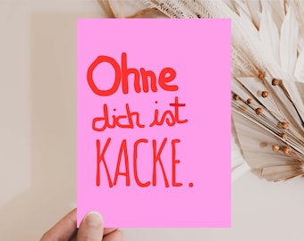 May include: A pink card with red handwritten text that reads "Ohne dich ist KACKE." The card is held by a hand, with a neutral background and decorative dried plants.