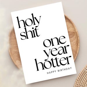 K&ouml;nnte beinhalten: Eine wei&szlig;e Geburtstagskarte mit schwarzem Text. Auf der Karte steht "holy shit one year hotter HAPPY BIRTHAY". Sie liegt auf einem Holztablett, mit einem geflochtenen Untersetzer und einem beigen Hintergrund.