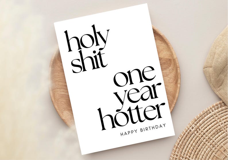 K&ouml;nnte beinhalten: Eine wei&szlig;e Geburtstagskarte mit schwarzem Text. Auf der Karte steht "holy shit one year hotter HAPPY BIRTHAY". Sie liegt auf einem Holztablett, mit einem geflochtenen Untersetzer und einem beigen Hintergrund.