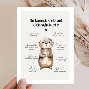 Puede incluir: Una tarjeta de felicitación con el texto "Du-kannst-stolz-auf dich-sein-Karte" y una ilustración de una linda nutria. La tarjeta tiene un fondo beige y presenta frases alentadoras en alemán. La tarjeta está sostenida por una persona.