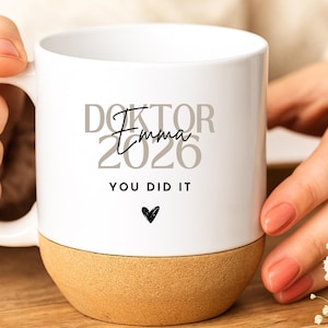 Pode incluir: Uma caneca de cerâmica branca com base de cortiça, apresentando o texto DOKTOR 2026 em cinzento, o nome Emma em letra cursiva preta e o texto YOU DID IT em letras maiúsculas pretas acima de um pequeno ícone de coração preto.