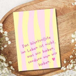 May include: A greeting card with a yellow and pink striped design. The card features the German text "Das Wertvollste im Leben ist nicht, was wir haben, sondern wen wir haben." A small pink heart is at the bottom.